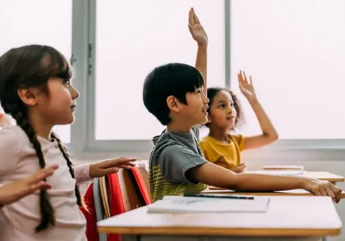 Two kids in class are raising their hands while a third kid is about to